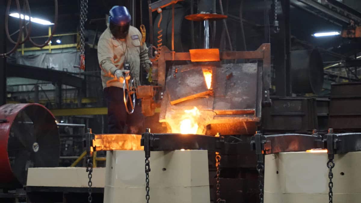 A Worker At Badger Foundry Is Pouring Molten Metal Into A Large Metal Container.