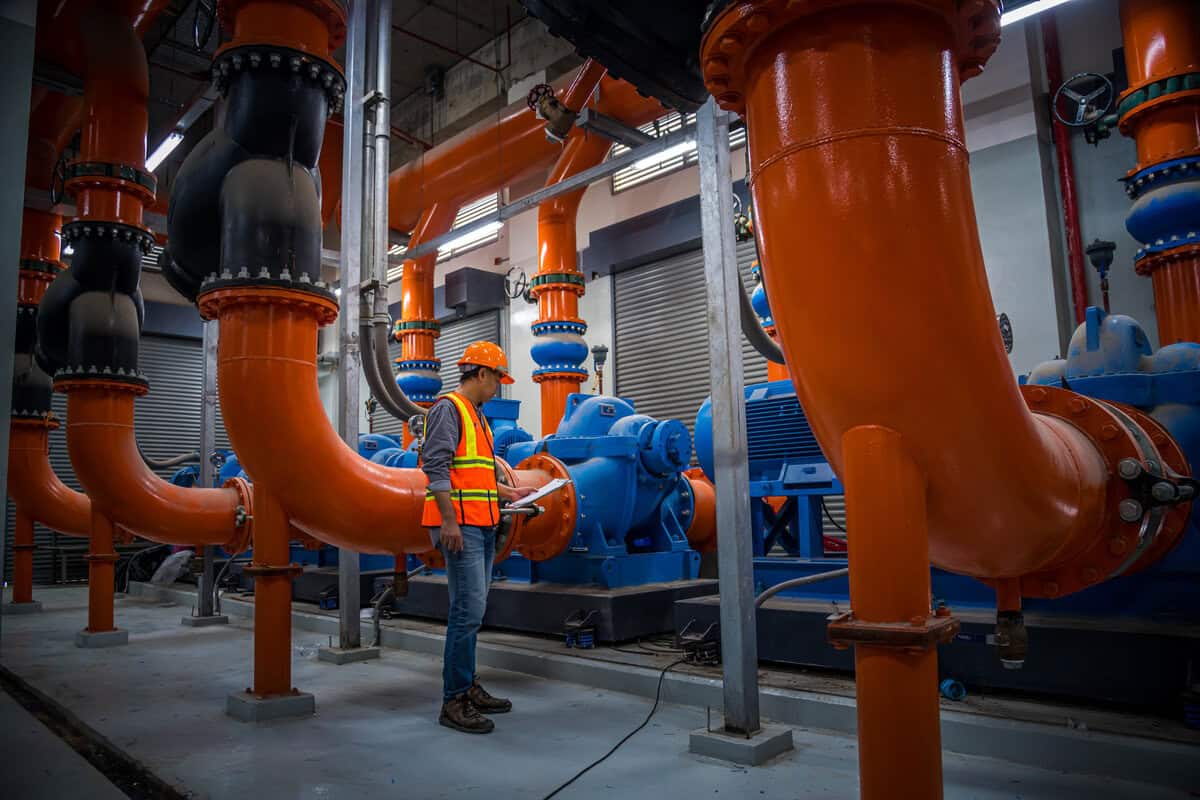 A Man In An Orange Vest Standing Next To A Large Pipe.